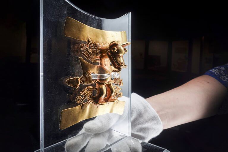 Gold figure mounted on a clear acrylic stand held in a gloved hand, lit with the Halumin H18 lighting device
