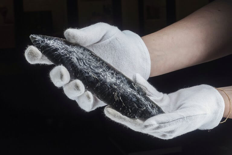 Museum curator holding an obsidian spear point in white cotton gloves, museum artifact photography with the Halumin H18