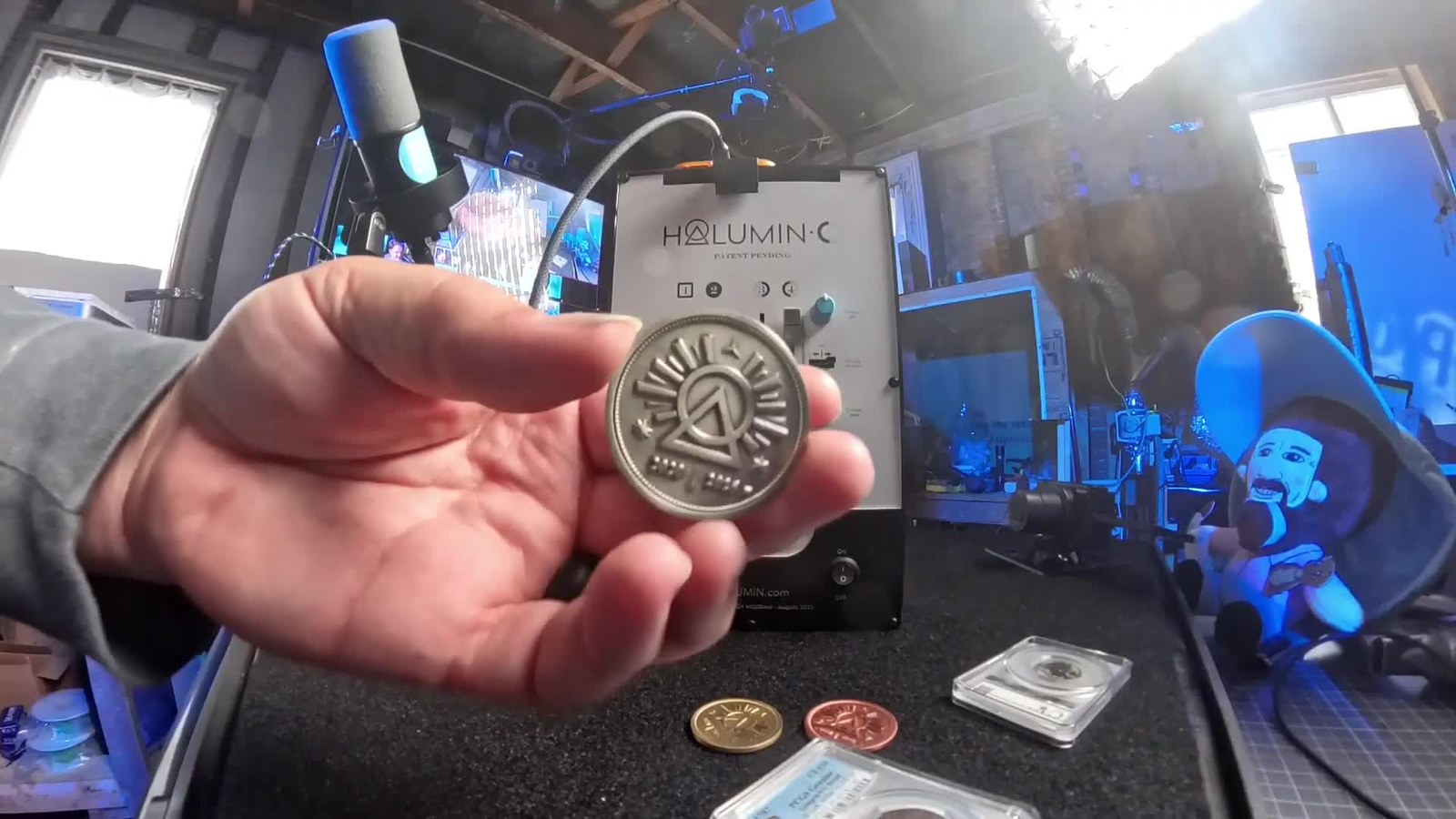 Holding the solid nickel Halumin coin, logo side up, in front of the Halumin-C, with two more Halumin coins and the slabbed coins on the table