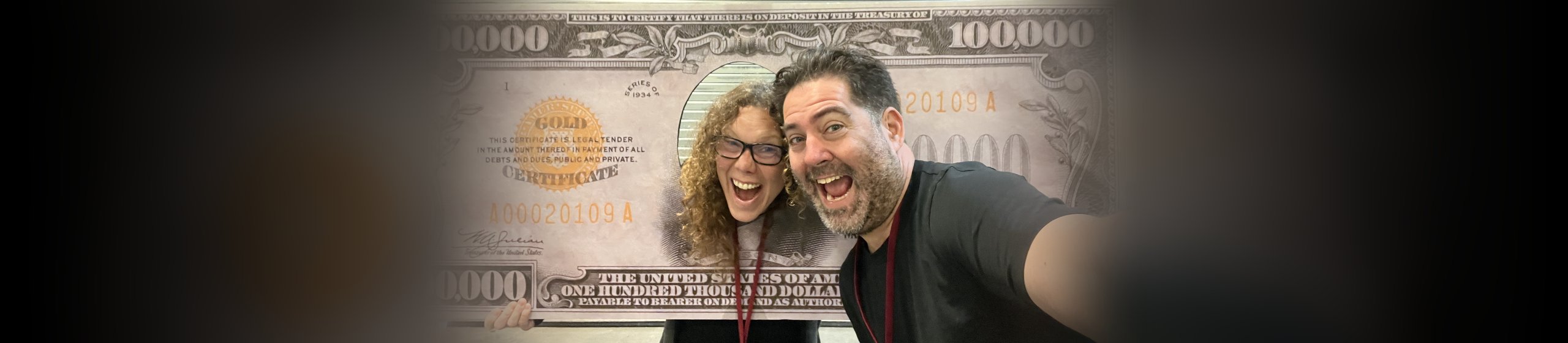 Halumin founder Jeremy and a team member grinning in a selfie with a giant $100,000 gold certificate prop at a coin show