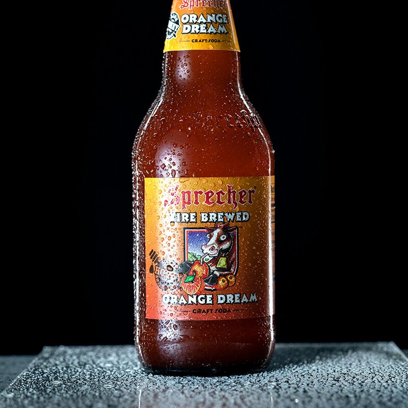 Sprecher Orange Dream soda bottle covered in water droplets on a wet surface, lit with Open Cylindrical™ lighting from the H18